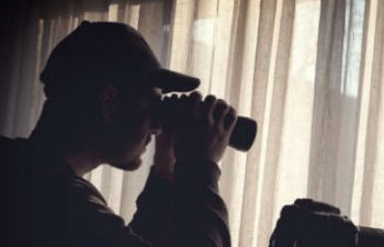 a man wearing a baseball cap using binoculars to observe an appartment in the other building