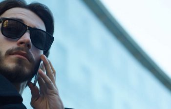 Beverly Hills private investigator in front of a building using a phone while observing the area through dark sunglasses
