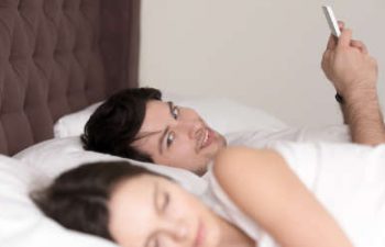 cheerful man in bed texting his lover and glancing at a sleeping by his side woman