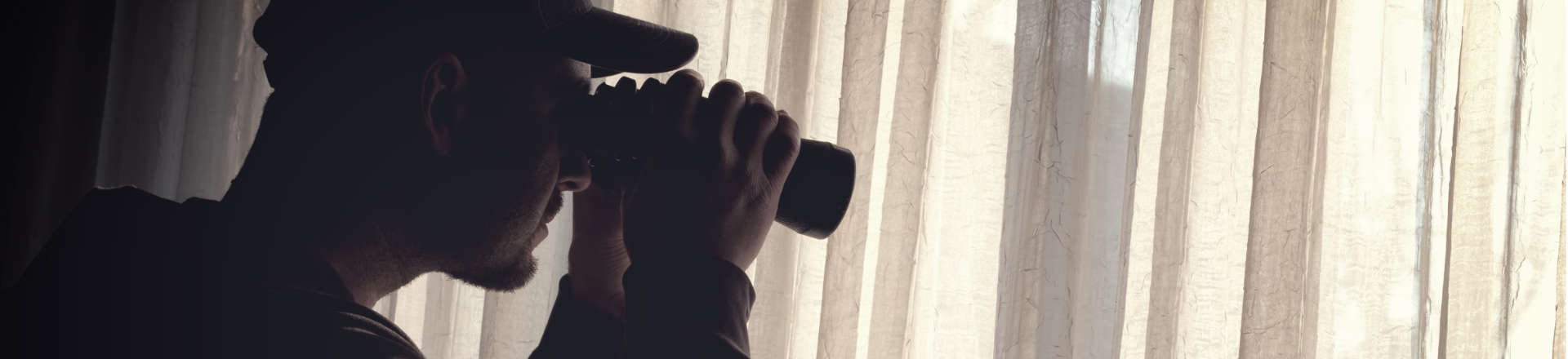 a man wearing a baseball cap using binoculars to observe an appartment in the other building