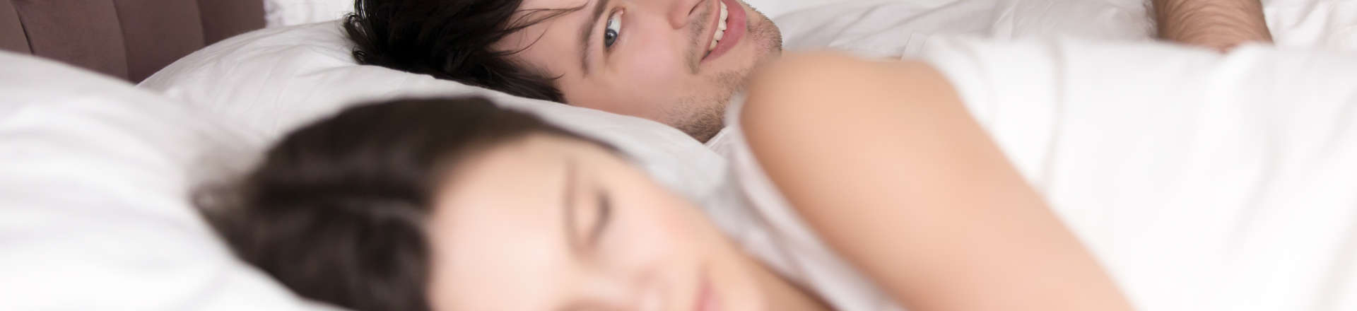 cheerful man in bed texting his lover and glancing at a sleeping by his side woman