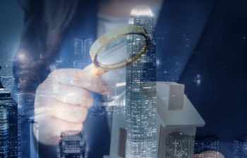 A blurred person holds a magnifying glass over a model building, with a cityscape in the background.