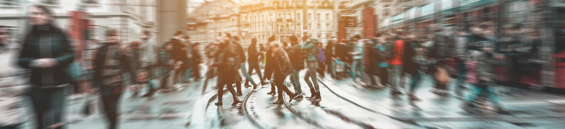 A busy street scene with many people walking in various directions, creating a sense of motion blur. The background includes buildings and a tram.