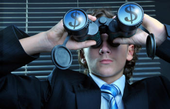 Person in a suit and tie looks through binoculars with dollar signs on the lenses, standing in front of window blinds.