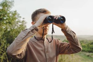 A man with modern binoculars against the sky and green forest.