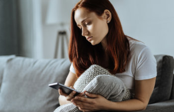 A woman with red hair sits on a grey couch holding a blanket and looking at her smartphone.