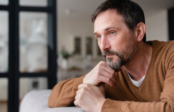 A man with a brown sweater looks pensively into the distance, resting his chin on his hand, in a softly lit interior space.