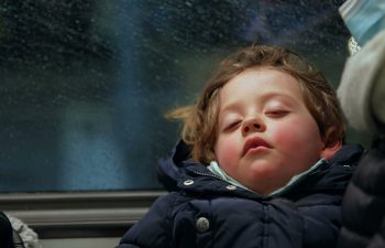 A young child with flushed cheeks sleeps against a window on public transportation, dressed in a dark winter coat, with raindrops visible on the glass.