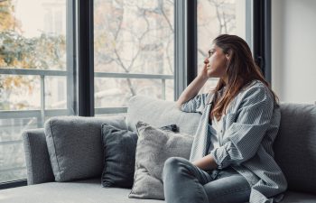 A woman sits on a gray couch by a window, resting her head on her hand and looking outside with a thoughtful expression.