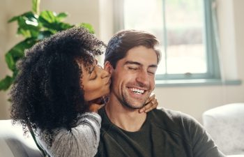 Happy man, woman and kiss on cheek on sofa for love, affection and proud for engagement. Interracial couple, hug and commitment in relationship with good news, trust and support at home with care